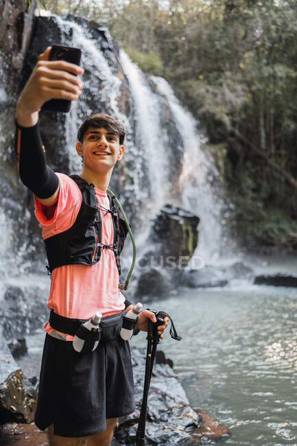 Escursionista maschio sorridente scattare auto sparato su smartphone mentre in piedi sullo sfondo della cascata e del lago nel bosco durante il trekking — Foto stock