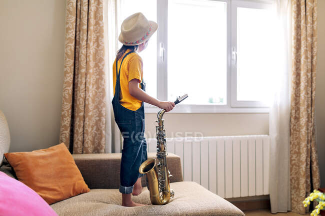 Back view of anonymous thoughtful barefoot child in hat with saxophone standing on sofa against window at home — Stock Photo