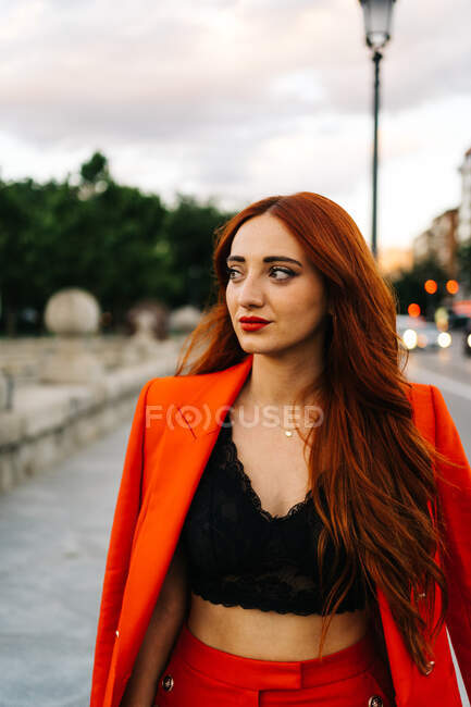 Charmante femelle aux longs cheveux roux et en costume orange tendance debout dans la rue le soir et regardant ailleurs — Photo de stock