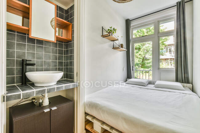 Interno di piccola camera da letto luminosa con letto pallet situato vicino alla finestra e lavandino in appartamento contemporaneo — Foto stock