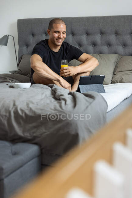 Deleitado macho sentado en la cama con un vaso de jugo de naranja y tableta de navegación mientras desayunaba en casa - foto de stock