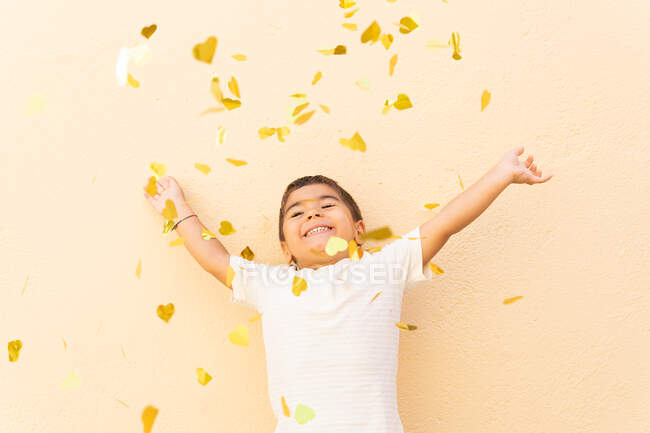Niño feliz en camiseta blanca de pie con los brazos levantados mientras vomita una pila de confeti amarillo escuchar en la pared naranja claro - foto de stock