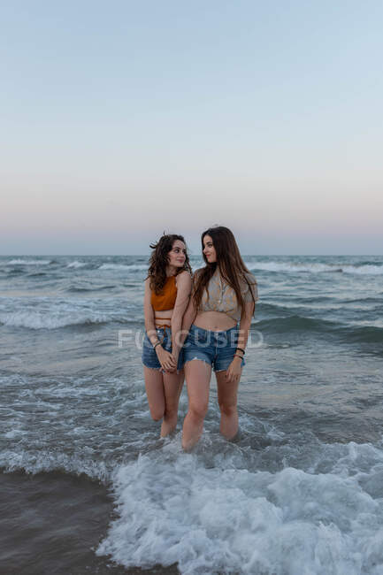 Junge Frauen halten Händchen, während sie beim romantischen Date in Meereswellen vor wolkenlosem Abendhimmel stehen — Stockfoto