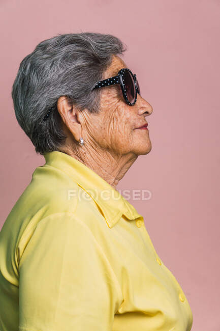 Vista lateral da mulher moderna feliz envelhecida com cabelos grisalhos e em óculos de sol na moda em fundo rosa em estúdio e olhando para longe — Fotografia de Stock