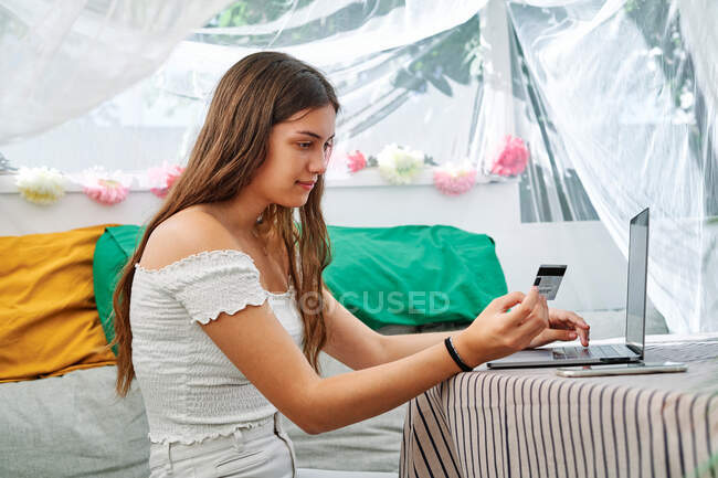 Délicieuse femme assise à table avec ordinateur portable et faisant des achats avec une carte en plastique pendant les achats en ligne dans la tente arrière-cour — Photo de stock