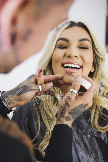 Cheerful blond woman smiling with closed eyes while tattooed makeup artist smearing liquid lipstick on lips of model — Stock Photo
