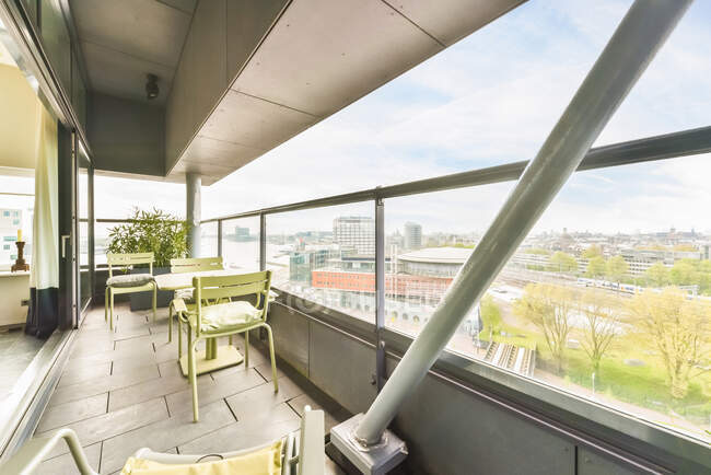 Стіл зі стільцями розташований на просторому балконі сучасної житлової будівлі зі скляним парканом, що переглядає міський пейзаж в літній день — стокове фото