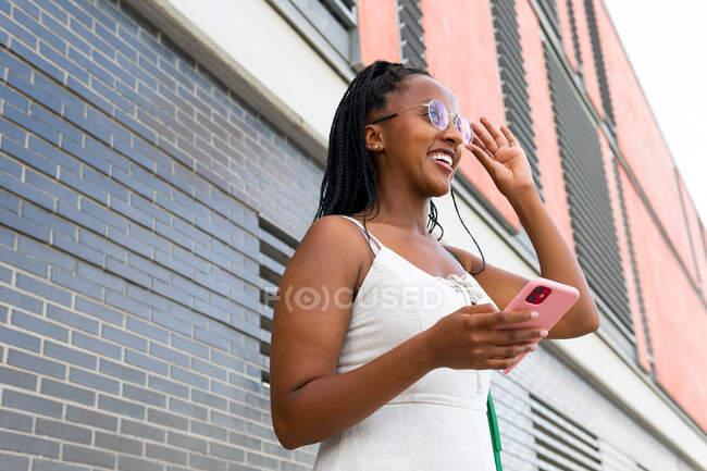 Низкий угол улыбки афроамериканской женщины с брадами в социальных сетях через мобильный телефон, стоя на улице со старыми зданиями в Барселоне — стоковое фото