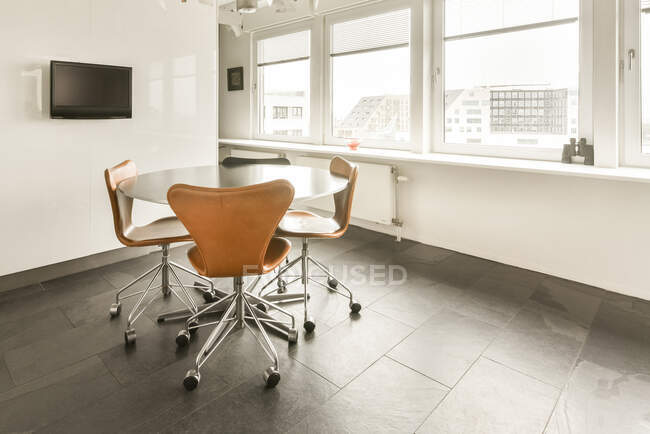 Mesa redonda y sillas situadas cerca de ventana panorámica en habitación espaciosa moderna con TV colgando en la pared blanca - foto de stock