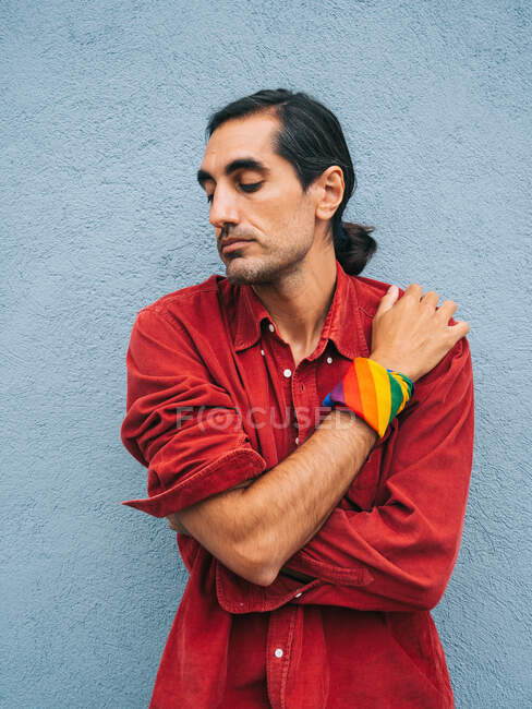 Heiterer ethnischer homosexueller Mann mit Regenbogenbandana auf der Hand, der sich zärtlich umarmt, während er vor dem Hintergrund einer grauen Mauer in der Stadt Selbstliebe ausdrückt — Stockfoto