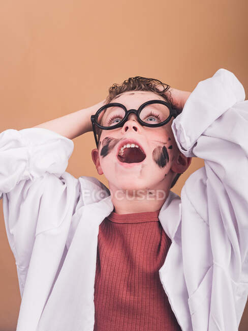 Chemiker-Junge im Labormantel und Plastikbrille auf beigem Hintergrund, die Hände auf dem Kopf — Stockfoto