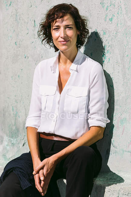 Задумчивая женщина в элегантной одежде стоит солнечный свет на потрепанной стене и смотрит в камеру — стоковое фото