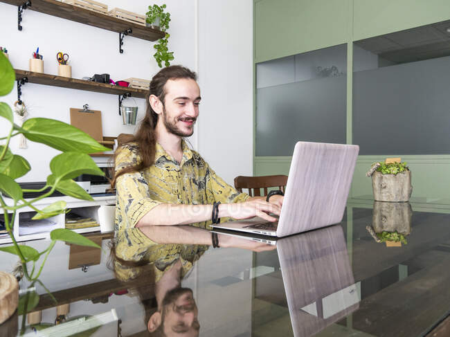 Entzückter Nacken mit langen Haaren tippt auf Netbook, während er an einem Projekt in einem modernen kreativen Arbeitsbereich arbeitet — Stockfoto