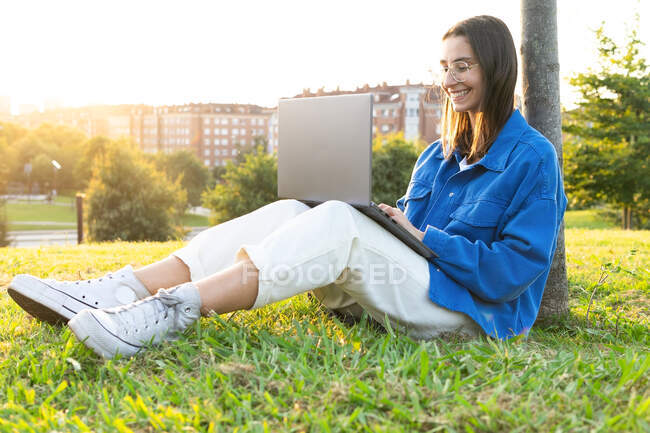 Side view of happy female freelancer leaning on tree and browsing netbook while working on project remotely in urban park on sunny day — Stock Photo