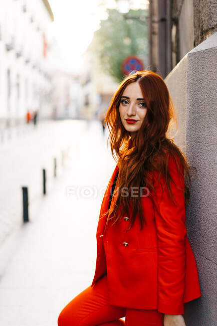 Side view of stylish female with ginger hair and in vivid orange suit leaning on wall of building in city street and looking at camera — Stock Photo