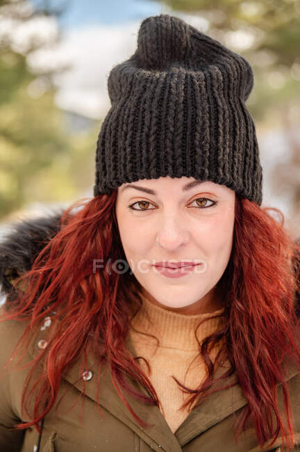 Cheerful female wearing warm hat and outerwear looking at camera in woods in winter — Stock Photo