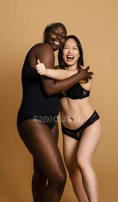 Entzückte multirassische weibliche Models in schwarzen Dessous, die sich auf beigem Hintergrund umarmen, um Körperpositivität im Studio zu begreifen — Stockfoto