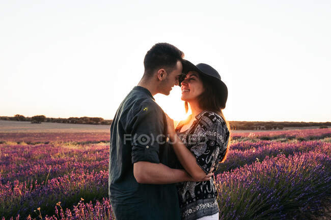 Visão lateral do homem de conteúdo abraçando namorada gentil no chapéu enquanto olha um para o outro contra a lavanda florescente na parte traseira iluminada — Fotografia de Stock