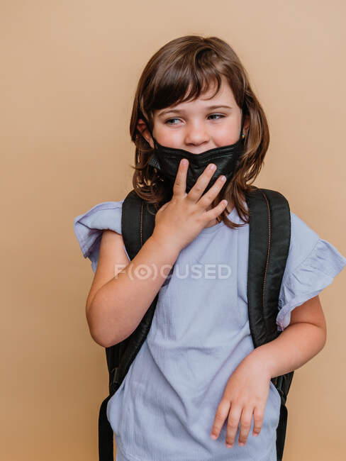 Delighted kid with rucksack and in protective mask from coronavirus on brown background in studio — Stock Photo