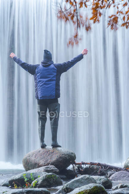Visão traseira de homem viajando em roupas quentes em pé sobre pedra com braços estendidos e admirando vista da rápida cachoeira em longa exposição no rio Lozoya no Parque Nacional de Guadarrama — Fotografia de Stock