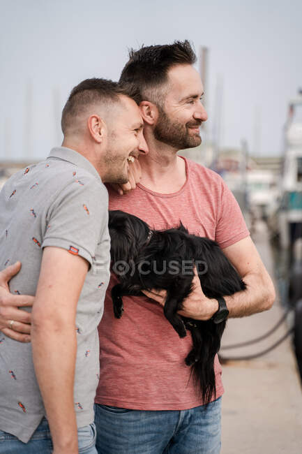 Вид сбоку веселых взрослых гомосексуальных мужчин с симпатичной собакой, смотрящих в гавань — стоковое фото