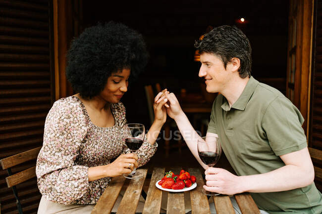 Vista lateral de alegre pareja multirracial cogida de la mano y con copas de vino tinto mientras disfruta de una cita romántica en la terraza de la casa a la mesa con plato de fresas - foto de stock