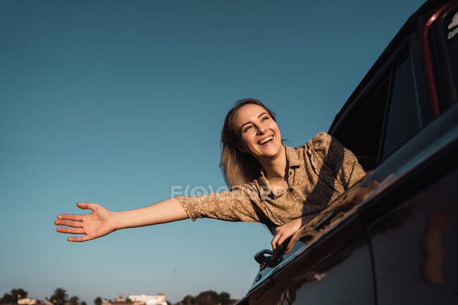 Low angle of cheerful female with outstretched arm sticking out of car window and enjoying freedom in summer evening — Stock Photo