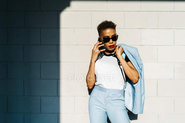 Stylish adult African American female with modern haircut and jacket conversing on cellphone against tiled wall with shadow in sunlight — Stock Photo
