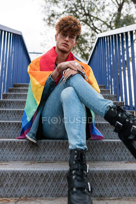Снизу стильный гей-мужчина с флагом ЛГБТ на плечах, сидящий на металлической лестнице в городе и смотрящий в камеру — стоковое фото