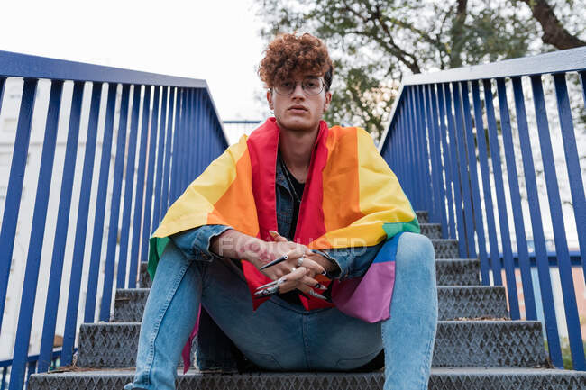Desde abajo de elegante gay masculino con bandera LGBT sobre hombros sentados en escaleras de metal en la ciudad y mirando a la cámara - foto de stock
