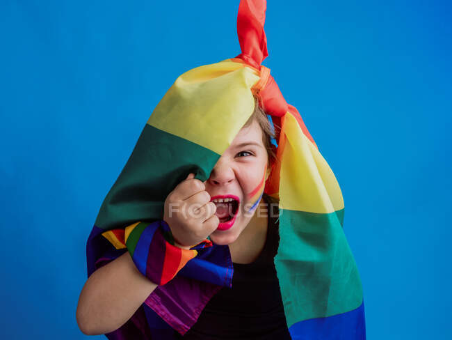 Linda chica con labios rojos y bandera del arco iris que cubre la mitad de la cabeza mirando a la cámara en el fondo azul mientras grita - foto de stock