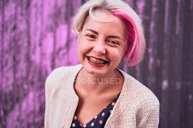 Encantadora mujer alternativa con el pelo teñido y en ropa de moda de pie contra la pared violeta en la ciudad y mirando a la cámara - foto de stock