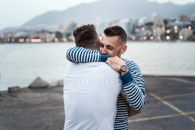 Hombre sincero abrazando pareja homosexual irreconocible mientras mira hacia otro lado contra el lago y el monte en la ciudad - foto de stock