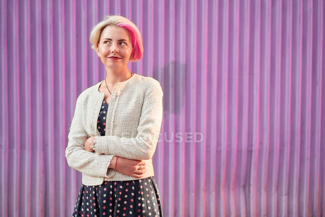 Очаровательная альтернативная женщина с окрашенными волосами и в модной одежде, стоящая напротив фиолетовой стены в городе и смотрящая в сторону — стоковое фото