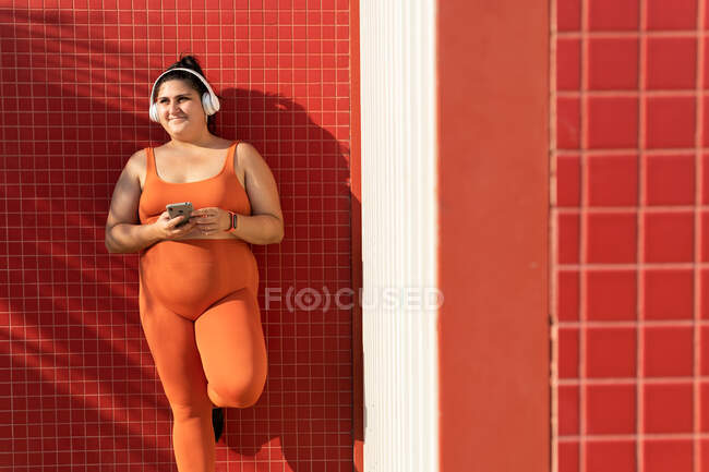 Cheerful ethnic female athlete with curvy body and cellphone listening to song from headphones while looking away against tiled wall — Stock Photo