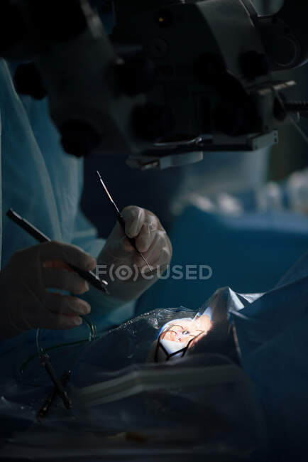 Cirujano ocular anónimo con instrumentos manuales operando al paciente en cama médica en el hospital sobre fondo borroso - foto de stock