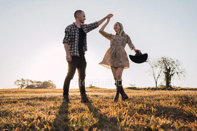 Von unten ein unbeschwertes Paar, das sich Händchen hält und an sonnigen Sommerabenden auf der Wiese tanzt, während es sich anschaut — Stockfoto