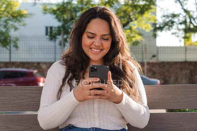 Веселая молодая латиноамериканская студентка просматривает мобильный телефон, отдыхая на деревянной скамейке на городской улице в летний день — стоковое фото