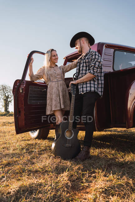 Fidanzato in cappello da cowboy con chitarra acustica mentre in piedi con la ragazza in rosso retro pick-up auto parcheggiata su strada sabbiosa in campagna — Foto stock