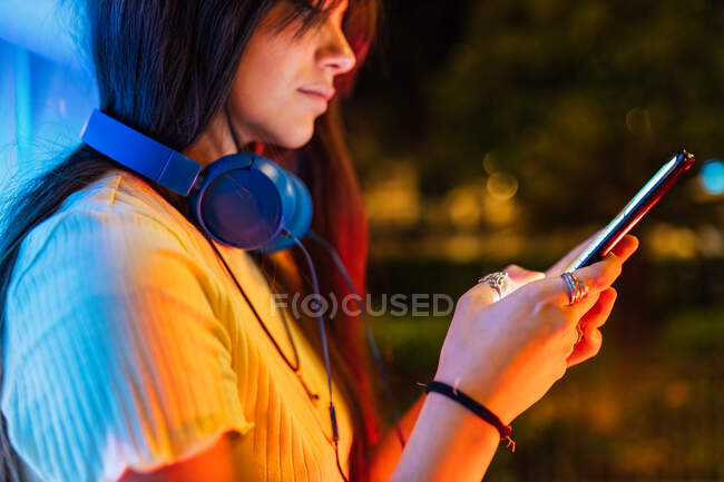 Side view of crop young female with headset text messaging on cellphone in night city — Stock Photo