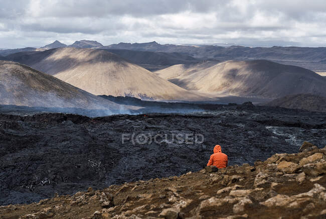 Rückansicht eines nicht wiederzuerkennenden Touristen, der den Fagradalsfjall bewundert, in dem sich Lava und Rauch zwischen Bergen unter wolkenverhangenem Himmel in Island ausbreiten — Stockfoto