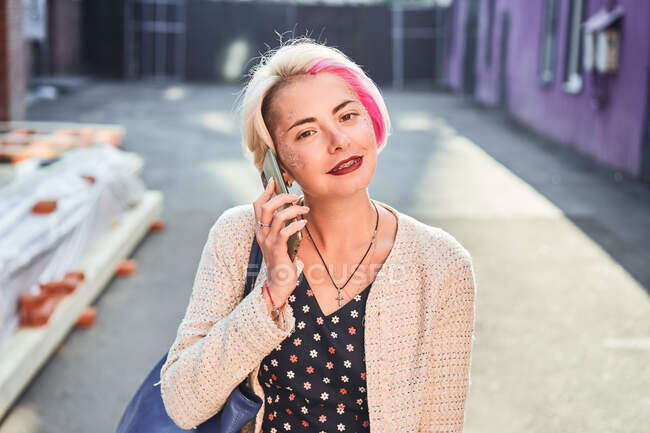 Cheerful alternative female with short hair standing in city and speaking on mobile phone — Stock Photo