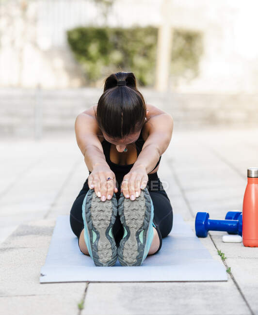Young female athlete in black sportswear doing warm up exercise while sitting on floor on city street — Stock Photo