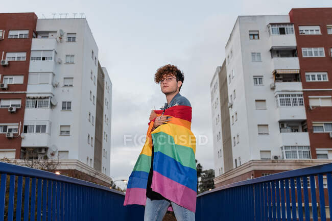 D'en bas d'un homme homosexuel sérieux debout avec drapeau arc-en-ciel sur le pont et regardant la caméra — Photo de stock