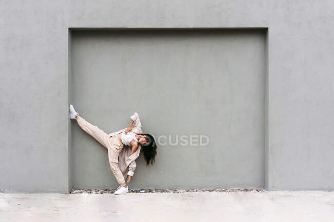 Hembra fresca creativa apoyada en la pared gris y bailando expresivamente en la calle de la ciudad - foto de stock