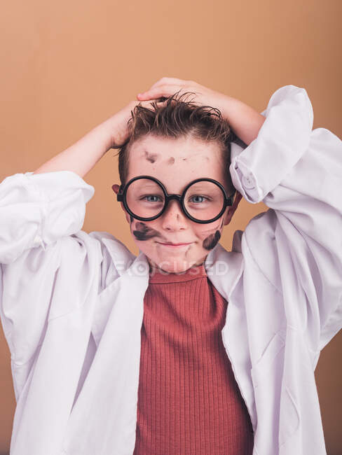 Chemist boy in laboratory robe and plastic glasses looking at camera on beige background with your hands on your head — Stock Photo