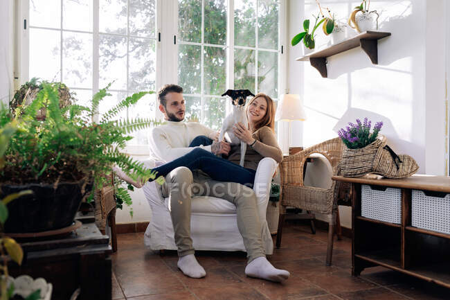 Бородатый мужчина с улыбающейся девушкой обнимает чистокровную собаку, отдыхая в кресле у окна в комнате — стоковое фото