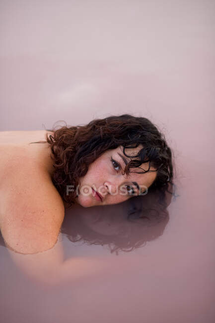 Tranquilo fêmea deitada com metade do rosto na água da lagoa rosa no verão e olhando para a câmera — Fotografia de Stock
