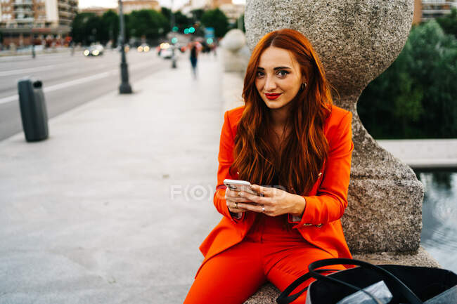 Happy stylish female with red hair and in vibrant orange suit sitting looking at camera on stone border in city and messaging on cellphone — Stock Photo