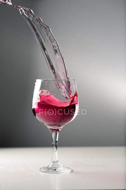 Cold alcohol red drink splashing out of glass goblet on grey background in studio — Stock Photo